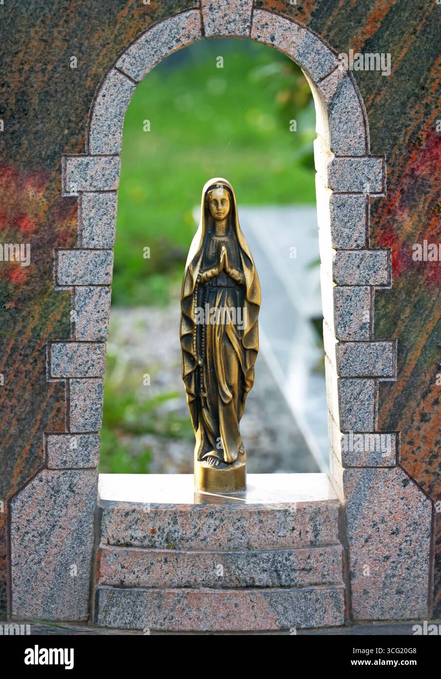 Petite statue de marie vierge dorée avec chapelet prie dans une niche de granit d'un cimetière Banque D'Images