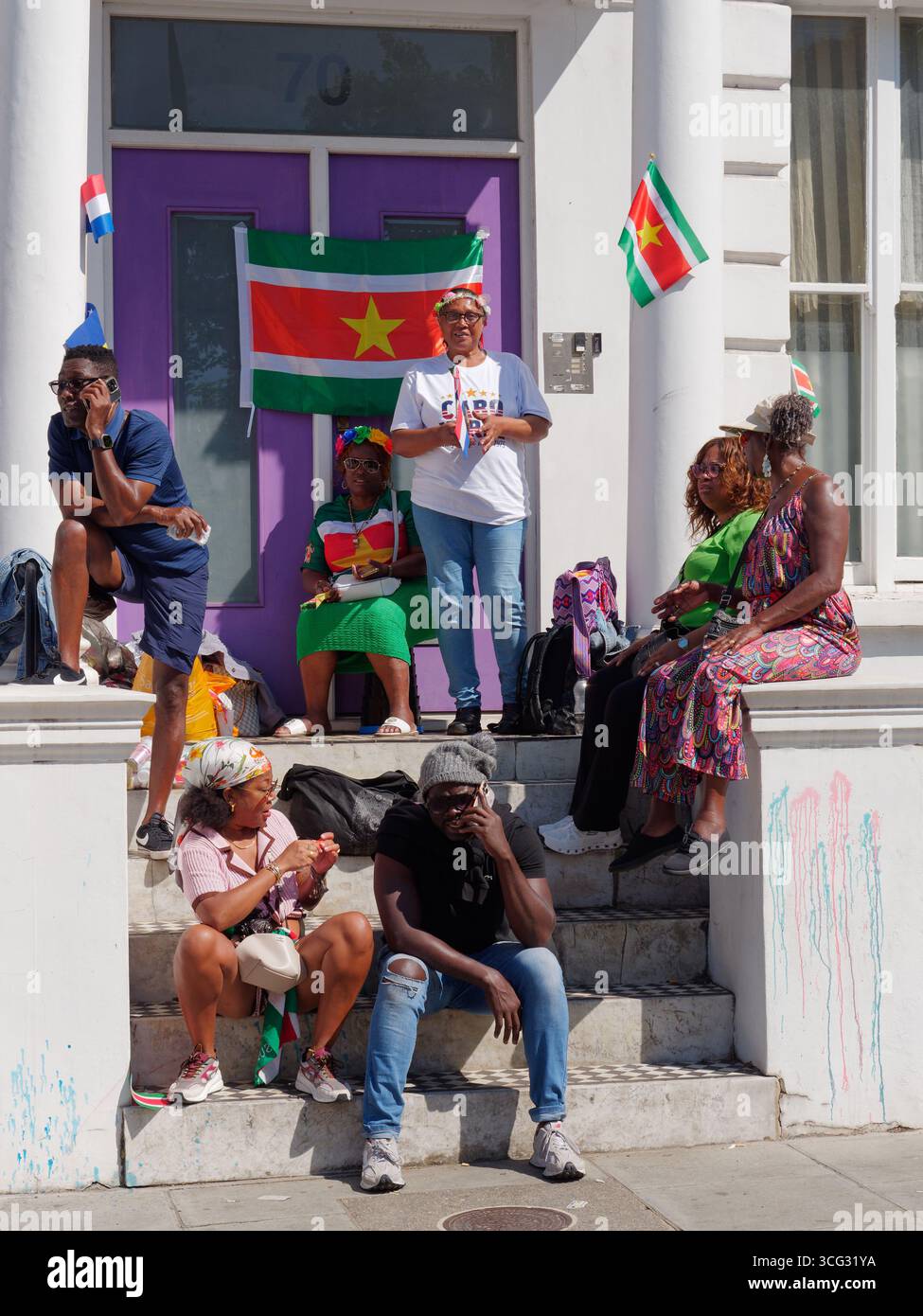 Des amis s'assoient sur les marches d'une propriété pour regarder le carnaval de Notting Hill, avec un drapeau drapé sur la porte d'entrée. Londres. Angleterre 25 août 2025 Banque D'Images