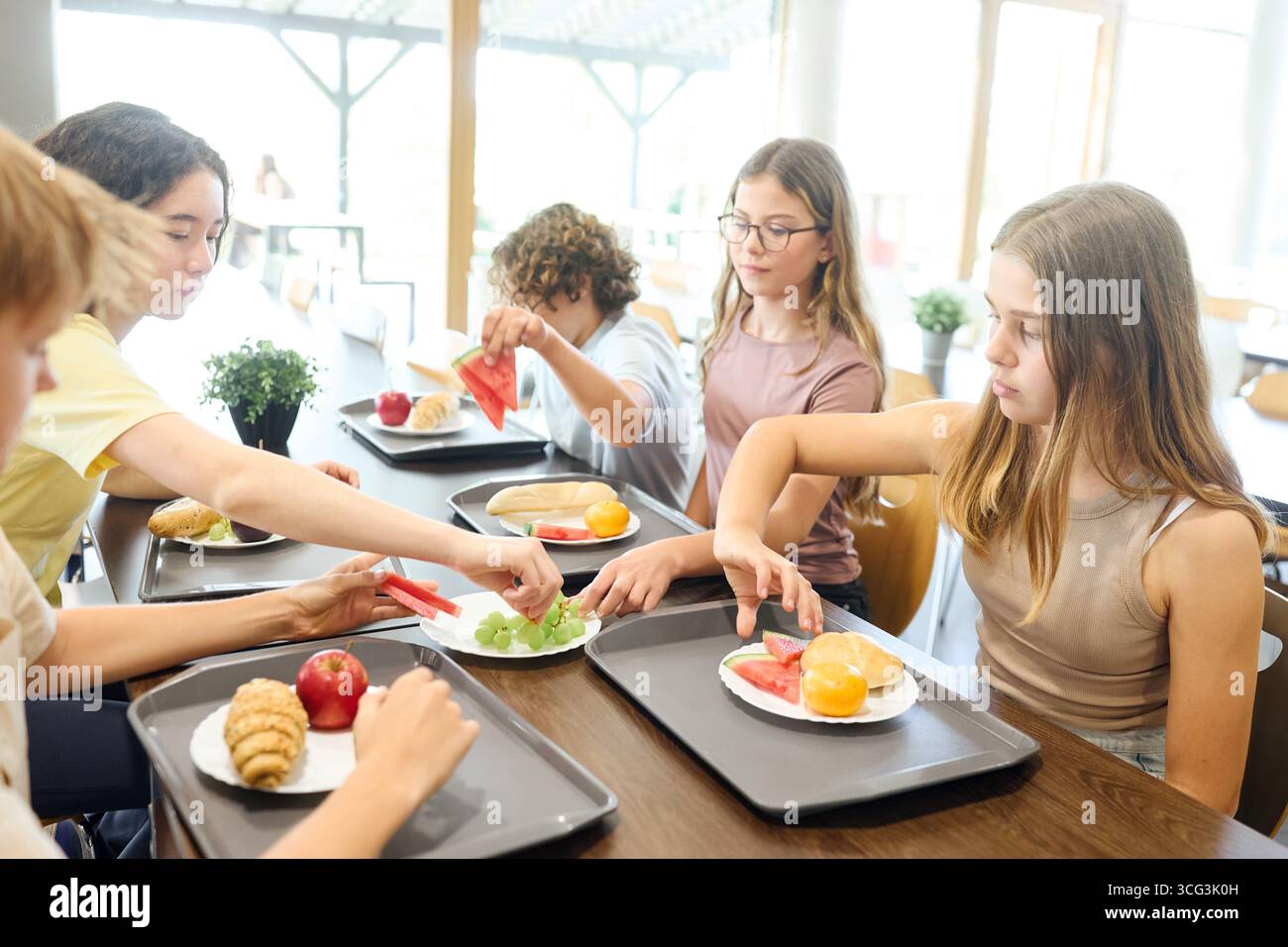 Les élèves profitent de leur déjeuner dans une cafétéria lumineuse de l'école, partagent des aliments frais et engagent une conversation autour d'une table commune. Un cheerfu Banque D'Images