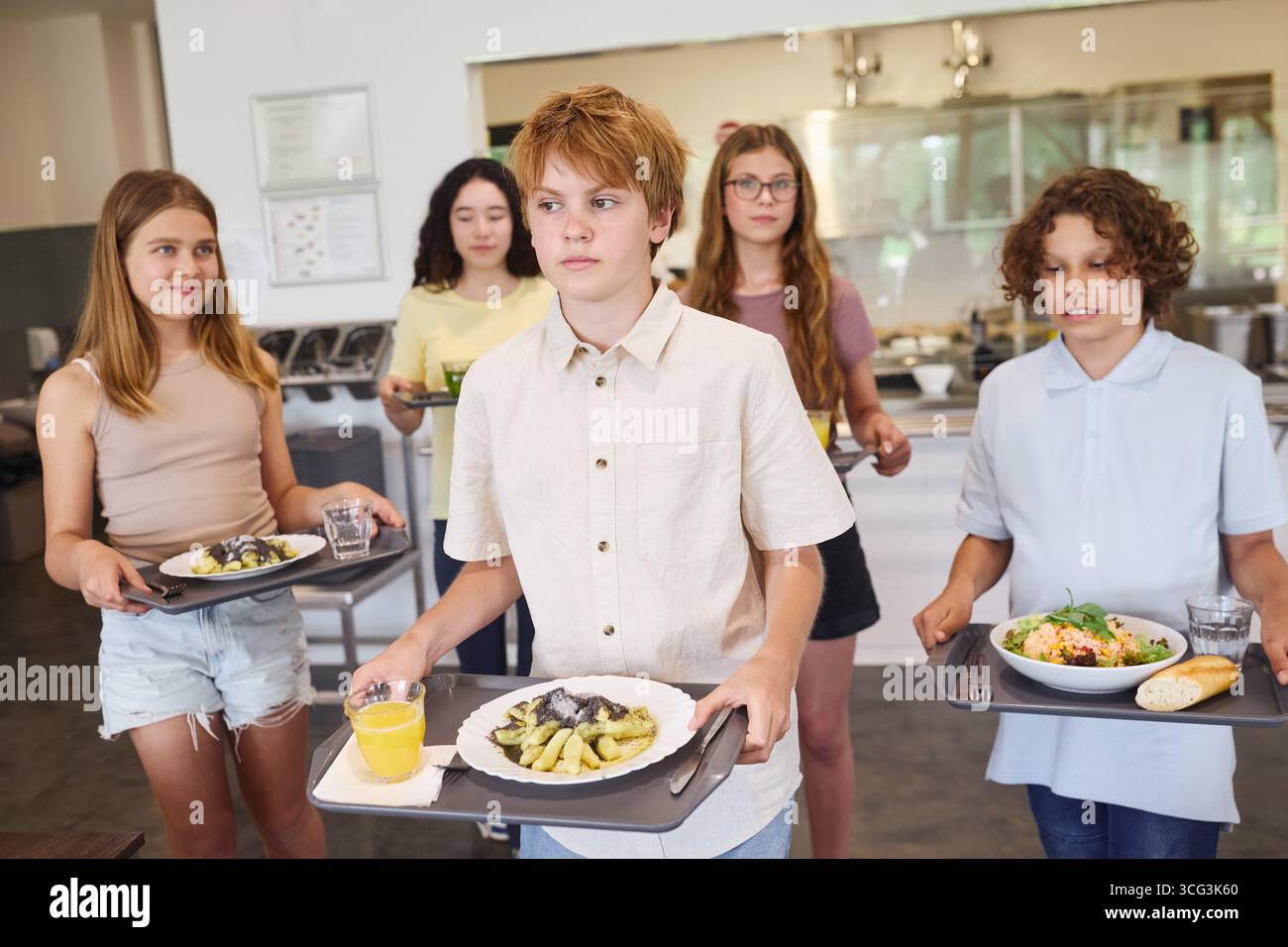 Un groupe diversifié d'étudiants transportant des plateaux avec des repas à travers une cafétéria d'école lumineuse. Banque D'Images