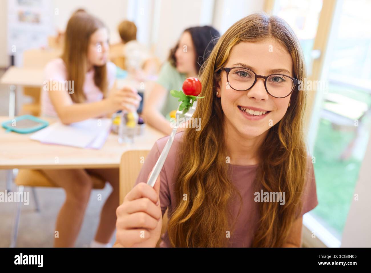 Une jeune fille joyeuse tient une fourchette avec une tomate, assise avec des amis dans une cafétéria de l'école. Le cadre dépeint un environnement vibrant et convivial, répondre Banque D'Images
