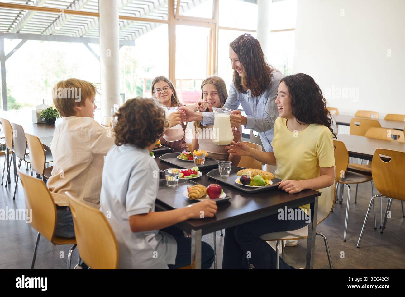 Un groupe d'élèves et un enseignant partagent le lait et la nourriture autour d'une table de cafétéria, démontrant des habitudes alimentaires saines et une interaction positive dans un edu Banque D'Images