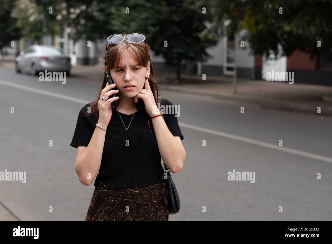 Une fille debout dans une rue de la ville a engagé une conversation téléphonique, montrant de l'inquiétude. Elle porte des vêtements décontractés et semble réfléchie ou inquiète Banque D'Images