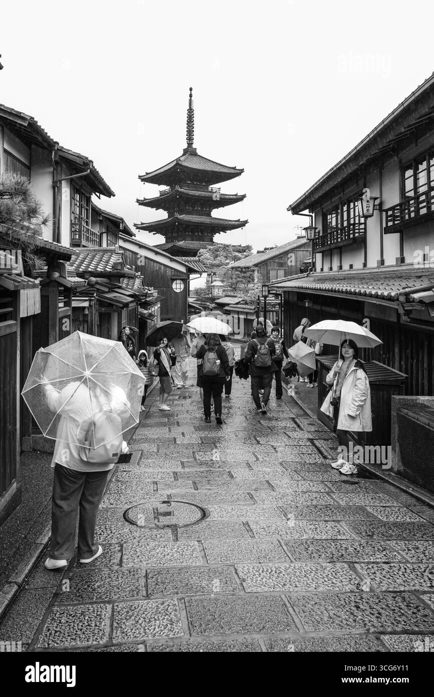 Une vue monochromatique du sentier Sannen-zaka sous le ciel couvert, avec des parapluies, des gens qui se promènent, et une pagode Yasaka frappante s'élevant dans l'arrière-pays Banque D'Images