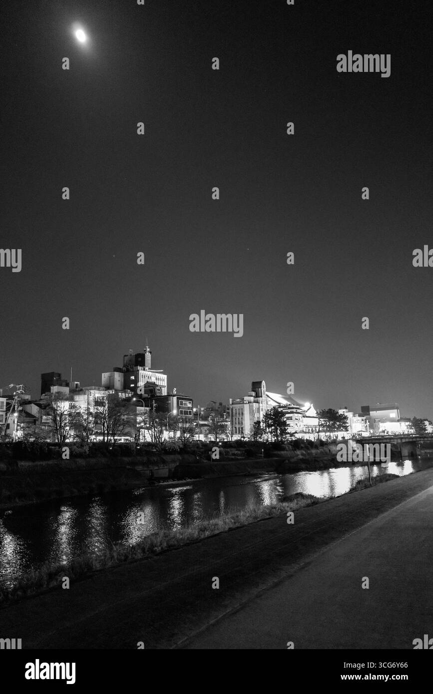 Une scène nocturne captivante en noir et blanc met en valeur une rivière Kamo calme reflétant les bâtiments illuminés de Kyoto et une lune rayonnante dans le ciel, évoquant Banque D'Images