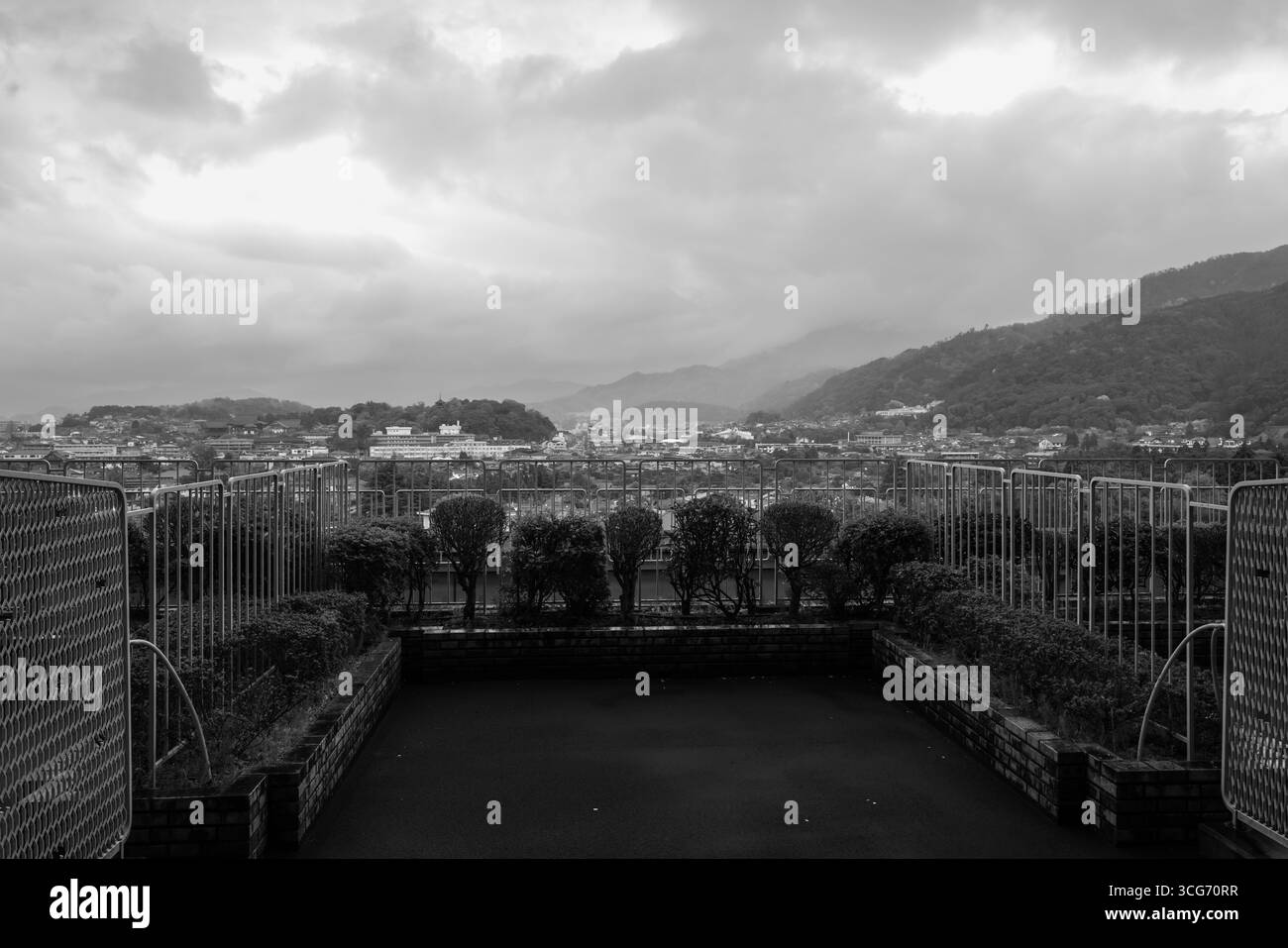 Une vue monochromatique capturant un paysage urbain encadré par des arbres et des clôtures, avec des montagnes brumeuses en arrière-plan, présentant un atmos serein et intemporel Banque D'Images