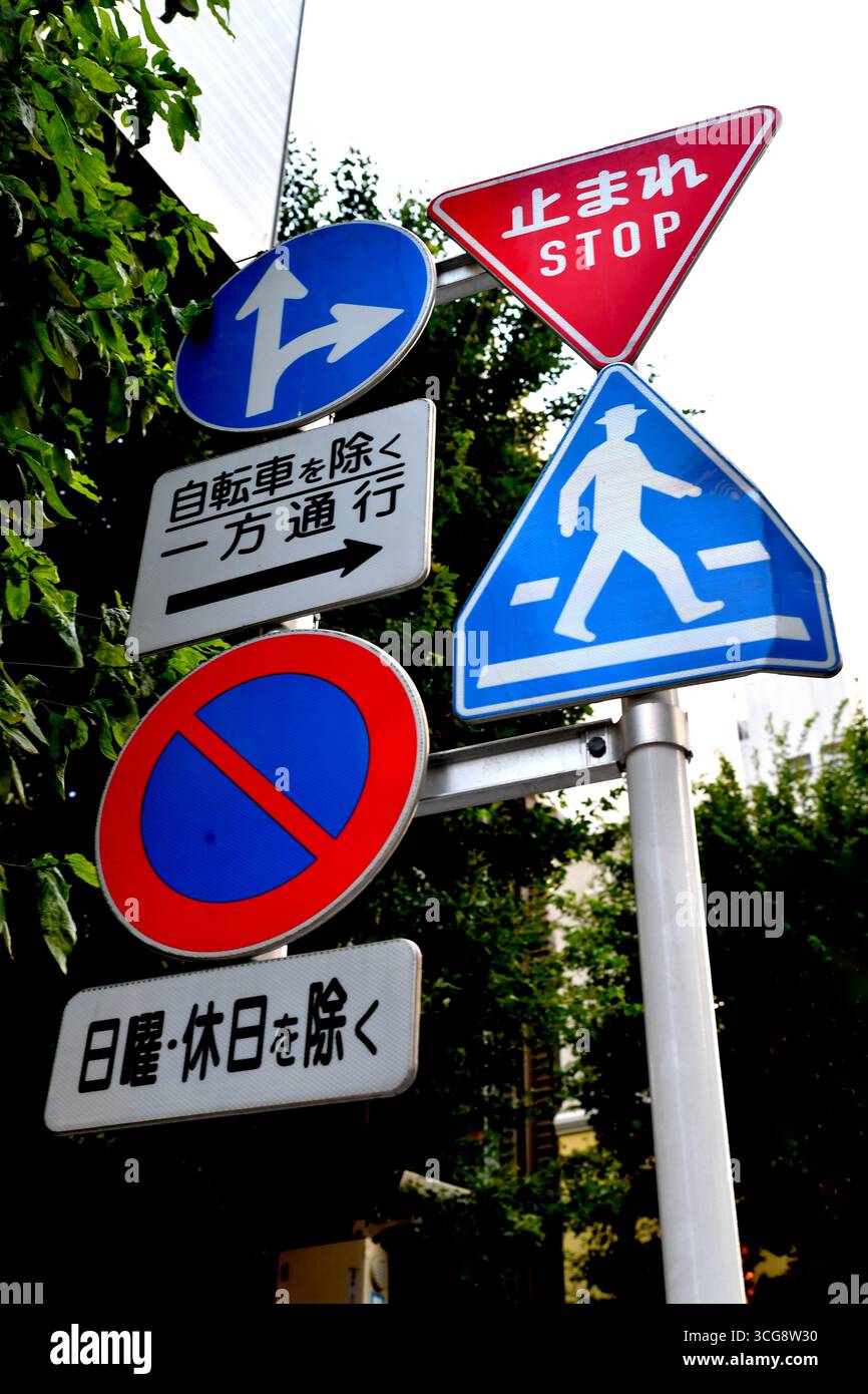 Signes de circulation japonais sur un poteau à Tokyo, incluant un panneau d'arrêt triangulaire rouge ("Stop" / "止まれ"), un panneau de passage piéton bleu à Tokyo - Japon Banque D'Images