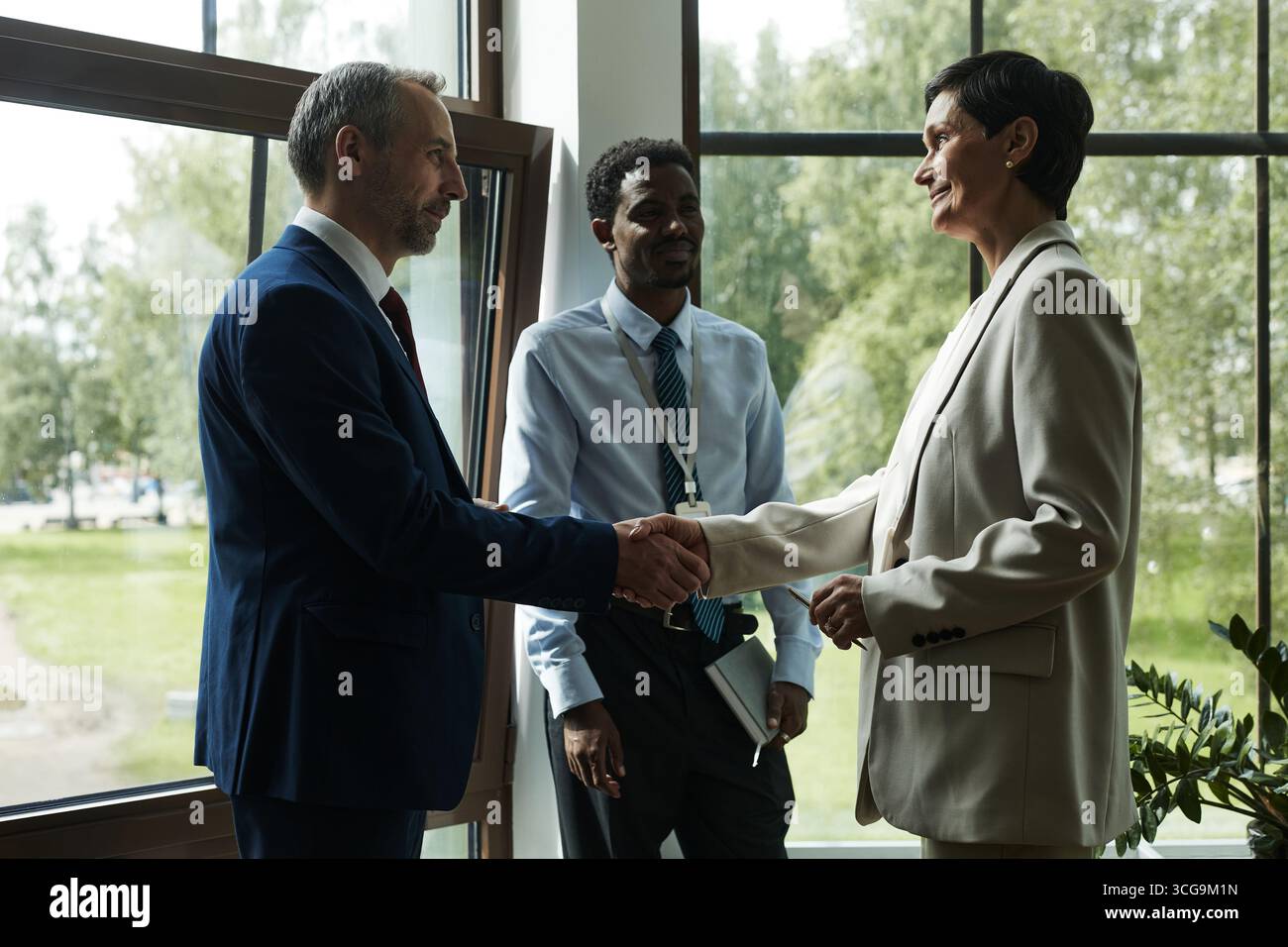 Homme d'âge moyen caucasien et collègue féminine en costume serrant la main, tandis que l'homme noir se tenant à proximité, observant l'interaction d'affaires, dans un bureau moderne avec de grandes fenêtres Banque D'Images