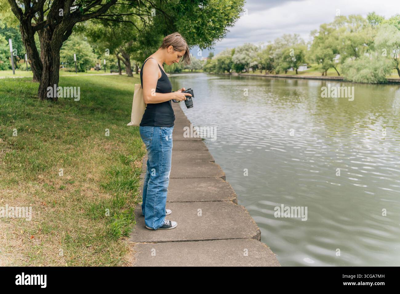 Femme des années 40 examinant des photos sur la caméra par la rive urbaine de la rivière avec sac fourre-tout Banque D'Images