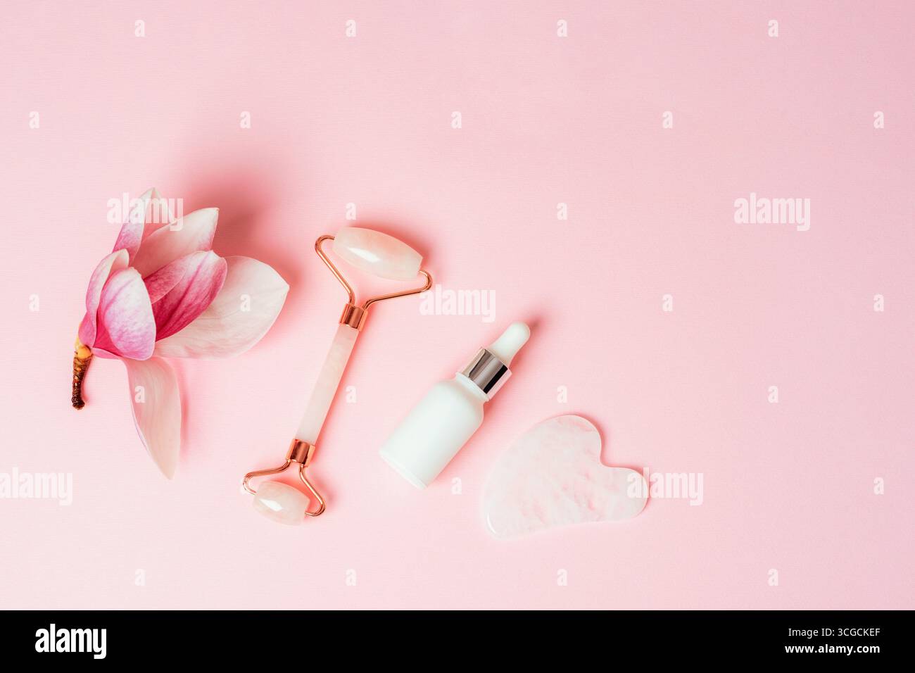 Rouleau facial en quartz rose, bouteille compte-gouttes de sérum blanc et pierre gua Sha avec fleur de magnolia sur fond rose. Concept de soins de la peau. Vue de dessus, pose à plat Banque D'Images