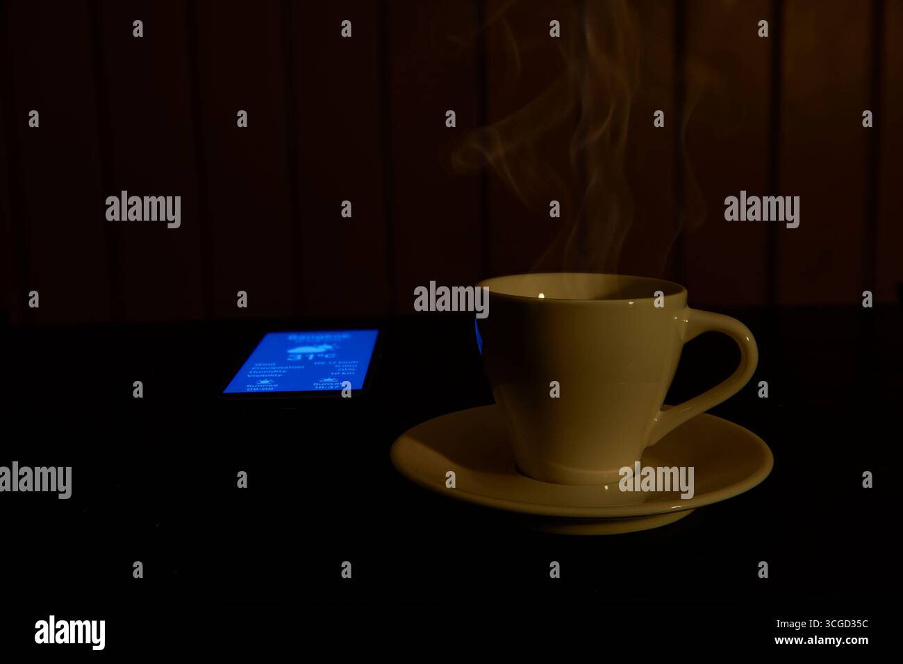 Une tasse de café chaude avec de la vapeur qui monte, éclairée par une lumière chaude, à côté d'un téléphone qui vérifie la météo. Un concept pour un matin confortable, slow living, rel Banque D'Images
