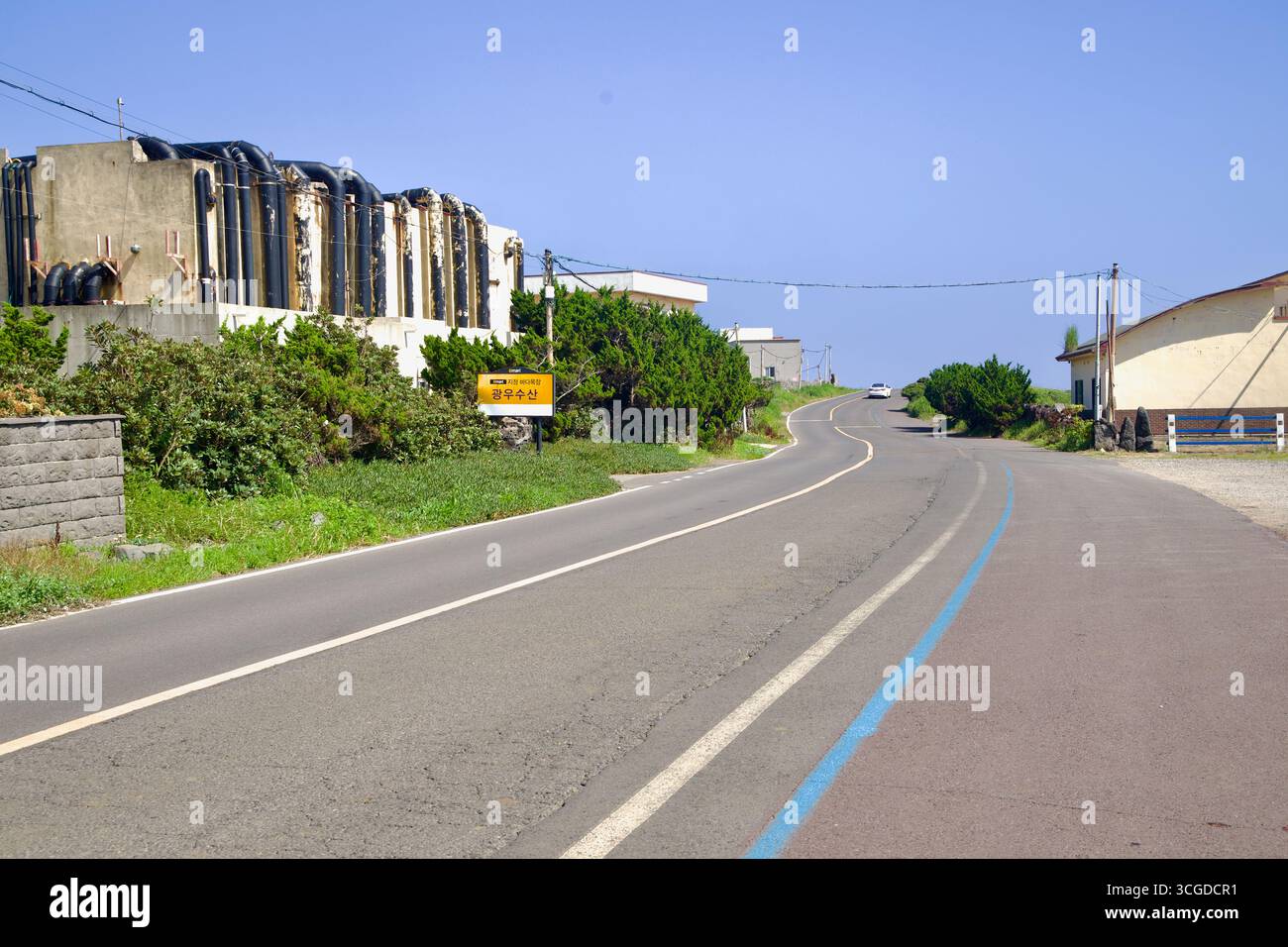 Yeongnak Coastal Road grimpe devant des growhouses d’ormeaux à bâches noires et une petite colline, avec une ligne de vélo bleue peinte guidant les cyclistes le long du wi de Jeju Banque D'Images