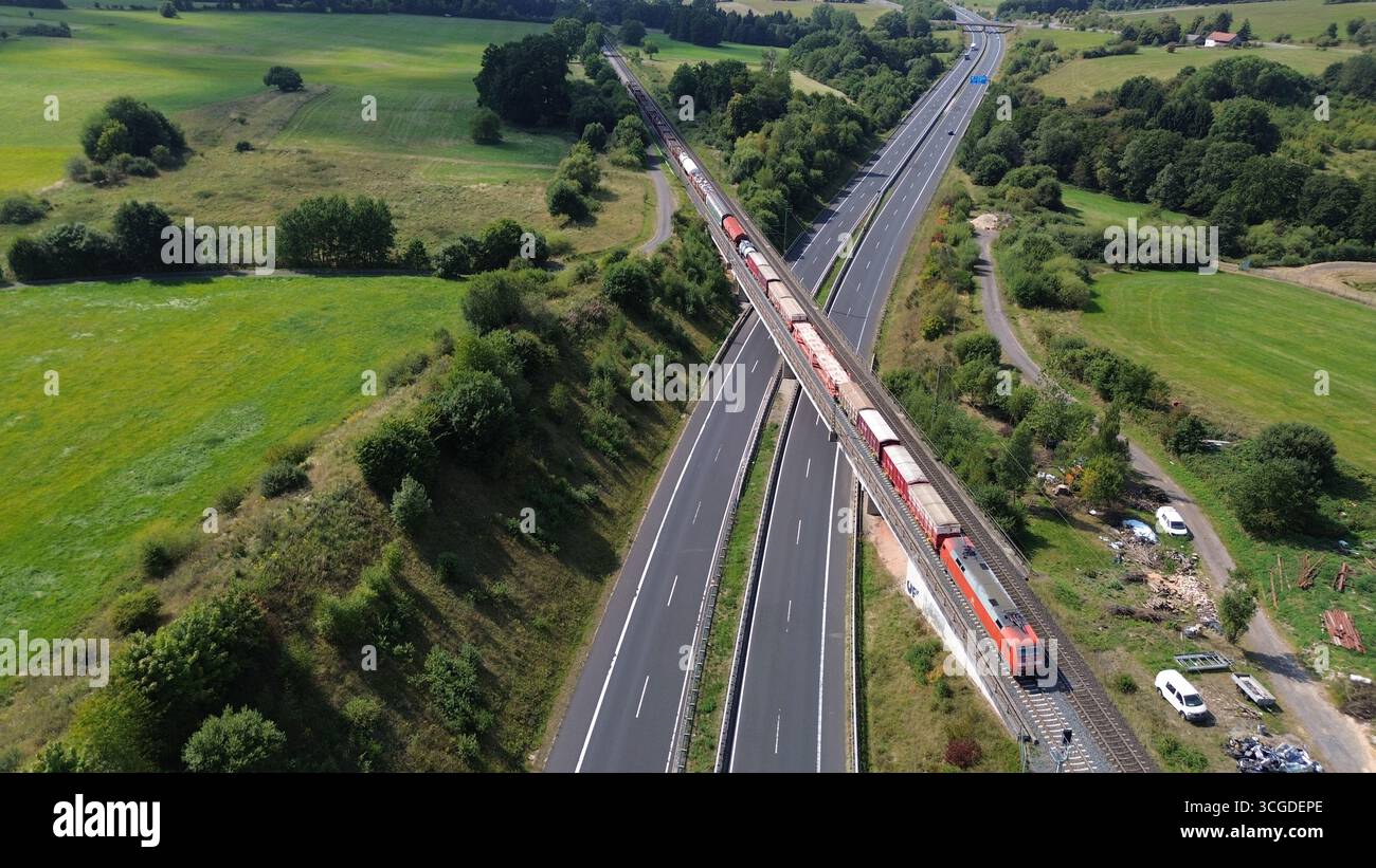 Vue aérienne du train de marchandises traversant le pont autoroutier à travers la campagne rurale avec des champs et de la forêt Banque D'Images