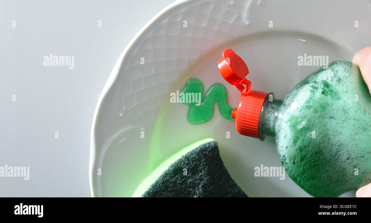 Verser le détergent à vaisselle d'une bouteille en plastique sur une assiette blanche avec un tampon à récurer sur un banc de cuisine blanc. Vue de dessus. Banque D'Images