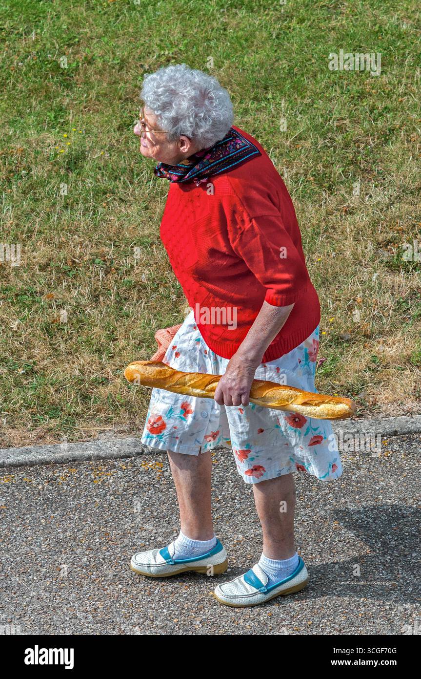Vue aérienne d'une femme âgée en pull rouge marchant sur le trottoir tenant une baguette - Preuilly-sur-Claise, Indre-et-Loire (37), France. Banque D'Images