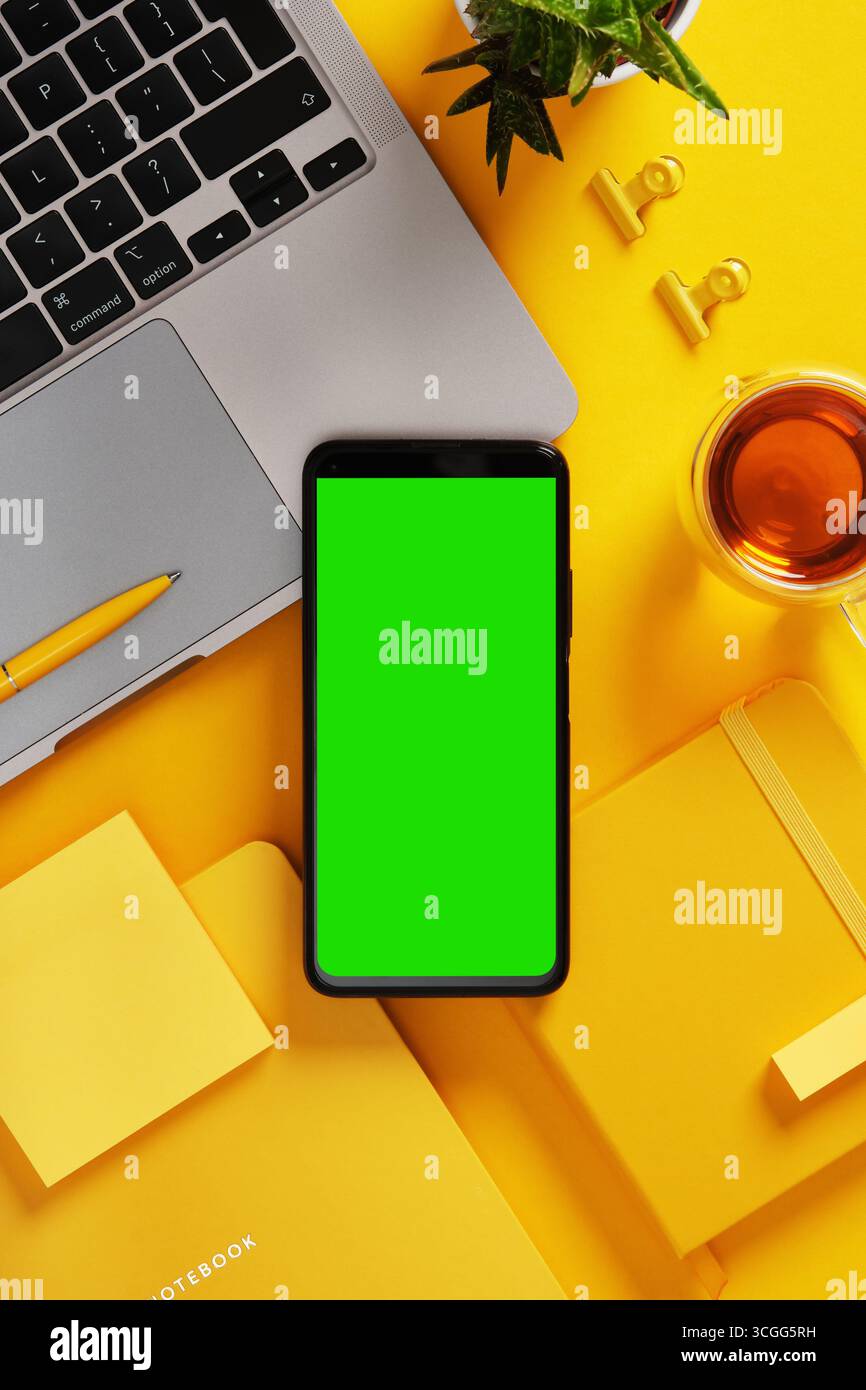 Smartphone avec écran vert près de l'ordinateur portable, stylo, cahiers, notes adhésives, plante, et du thé sur le bureau jaune. Présentez votre application, votre site Web ou votre capture d'écran. Banque D'Images