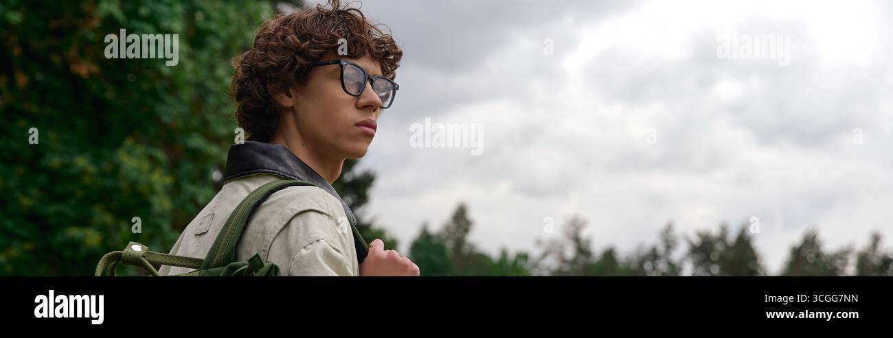 Un jeune homme aux cheveux bouclés et aux lunettes profite d'une promenade paisible en plein air parmi les arbres, réfléchissant. Banque D'Images