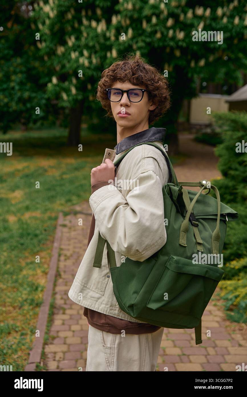 Beau jeune étudiant se promène dans un cadre extérieur tranquille avec un sac à dos à la main. Banque D'Images