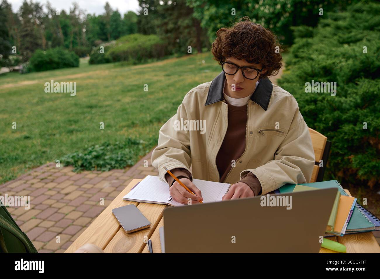 Un jeune homme étudie à une table en bois, concentré sur son ordinateur portable et ses livres au milieu de la verdure. Banque D'Images