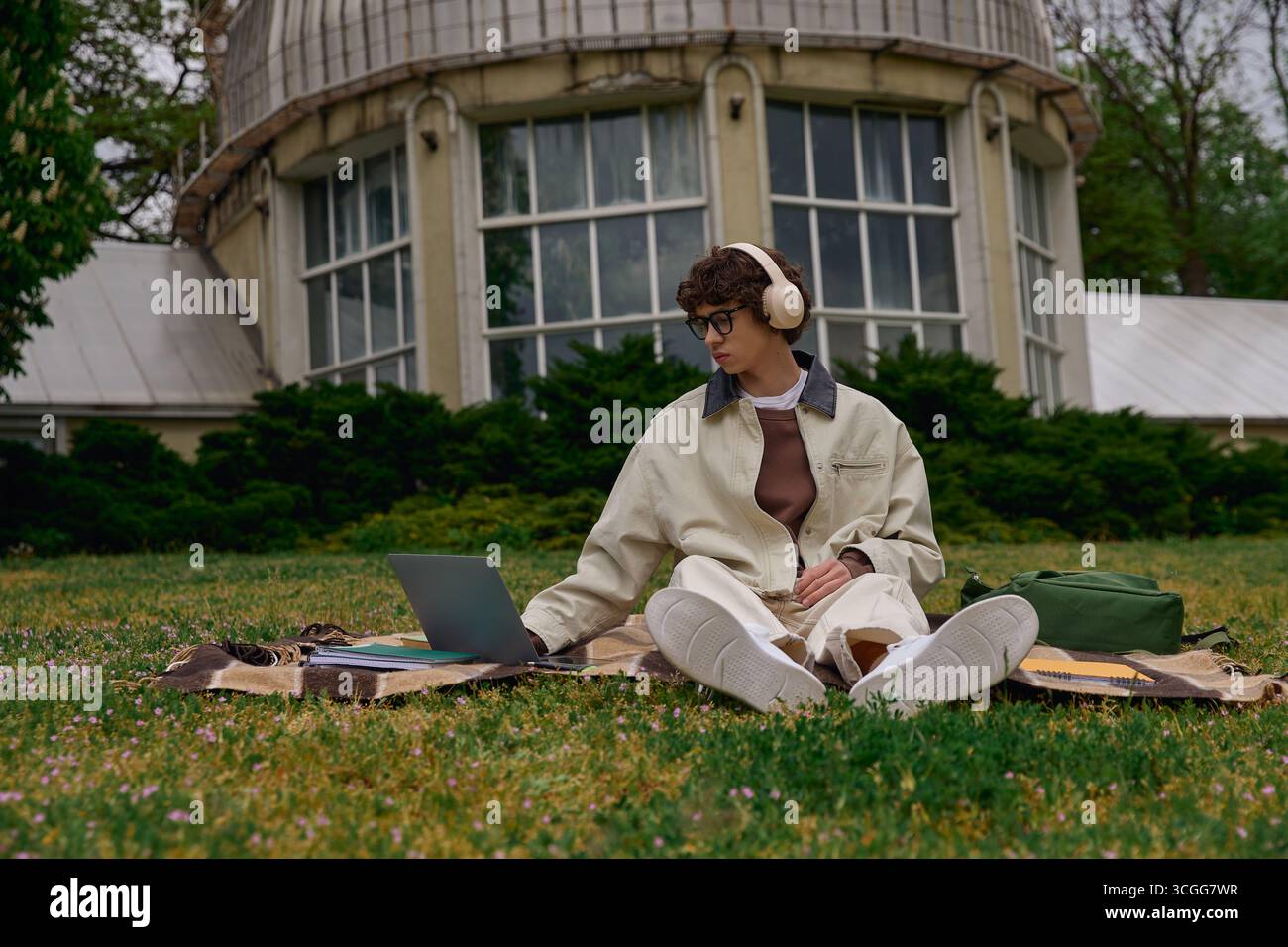 Un beau jeune étudiant est assis sur l'herbe, concentré sur son ordinateur portable tout en étant entouré de verdure. Banque D'Images