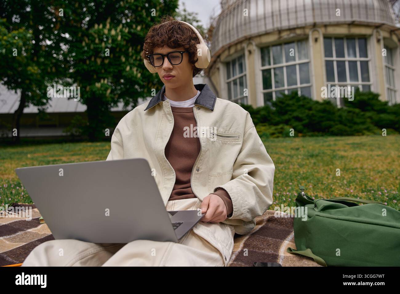 Beau jeune homme est assis sur l'herbe avec un ordinateur portable, axé sur les études dans un cadre extérieur tranquille. Banque D'Images