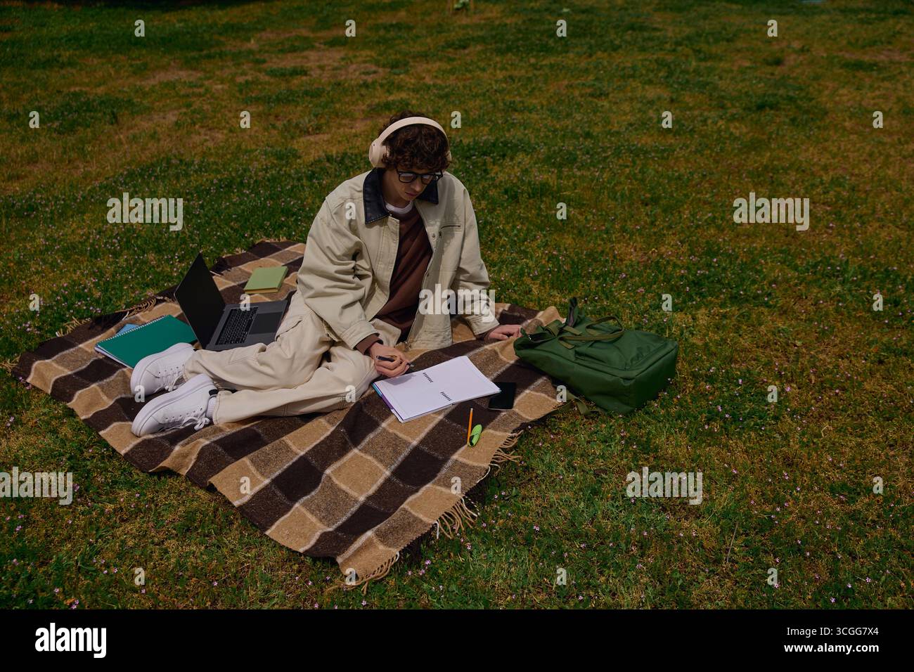 Jeune étudiant se détend sur une couverture, écrivant des notes tout en écoutant de la musique dans un parc verdoyant. Banque D'Images