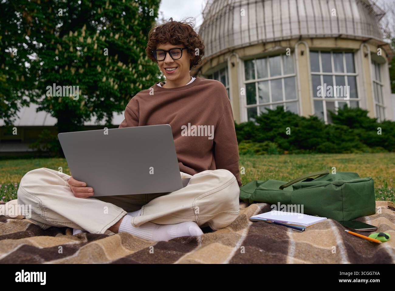Un jeune homme est assis confortablement sur une couverture, engagé avec son ordinateur portable dans un cadre magnifique en plein air. Banque D'Images