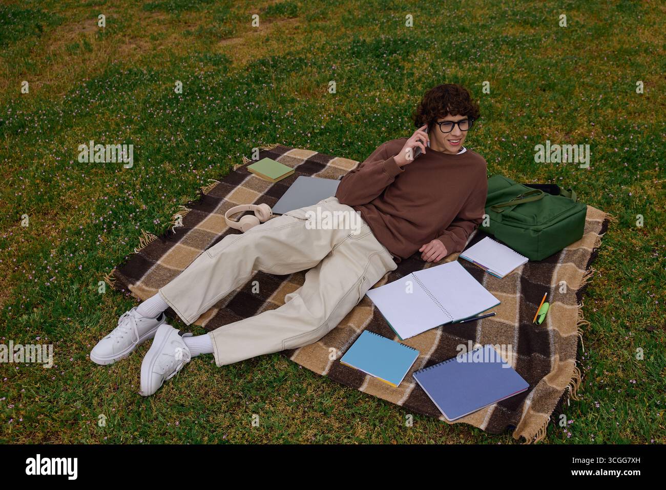 Un jeune homme se détend sur une couverture avec des cahiers et un sac à dos, étudiant dans la nature. Banque D'Images