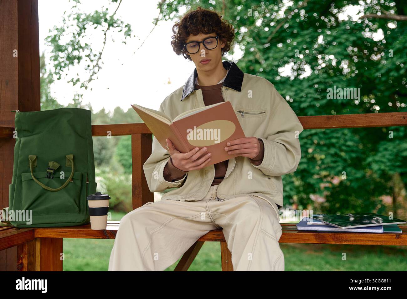 Un jeune homme est assis sur un banc, profondément engagé dans un livre au milieu d'arbres verts et d'une atmosphère sereine. Banque D'Images