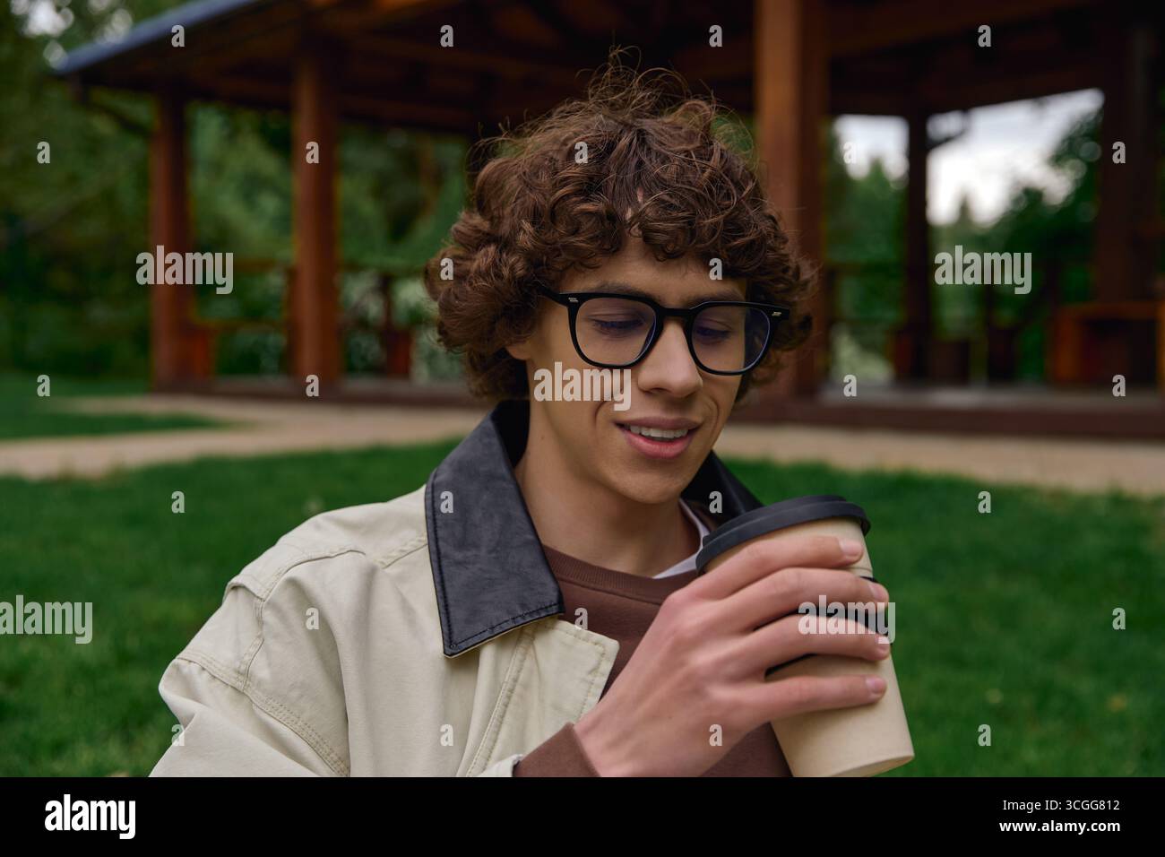 Un beau jeune étudiant se promène dans un cadre paisible en plein air, en savourant une boisson chaude. Banque D'Images