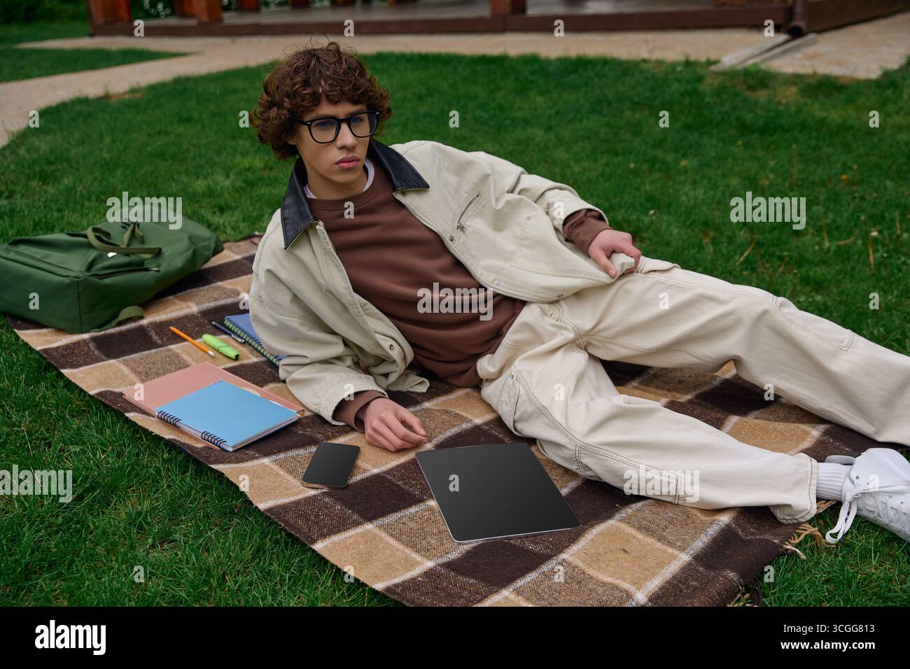 Relaxant en plein air, un beau jeune étudiant prend un moment pour se détendre avec du matériel d'étude sur l'herbe. Banque D'Images