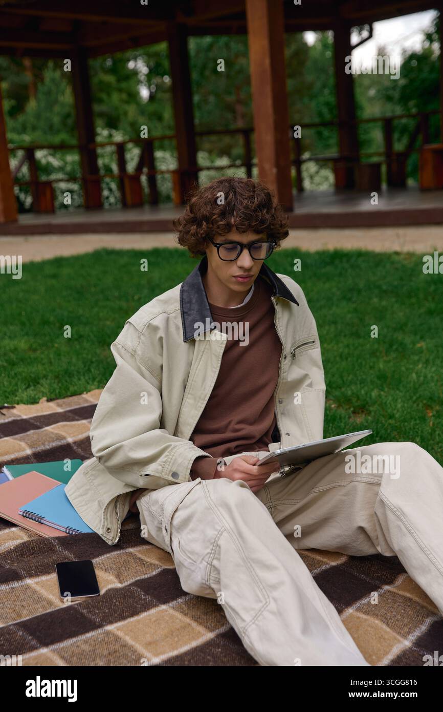 Un beau jeune homme est assis sur une couverture, lisant tranquillement dans un parc sous un gazebo par une journée ensoleillée. Banque D'Images