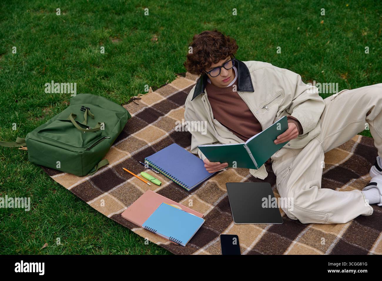 Un jeune homme se détend sur une couverture tout en lisant des livres dehors, entouré par la beauté de la nature. Banque D'Images