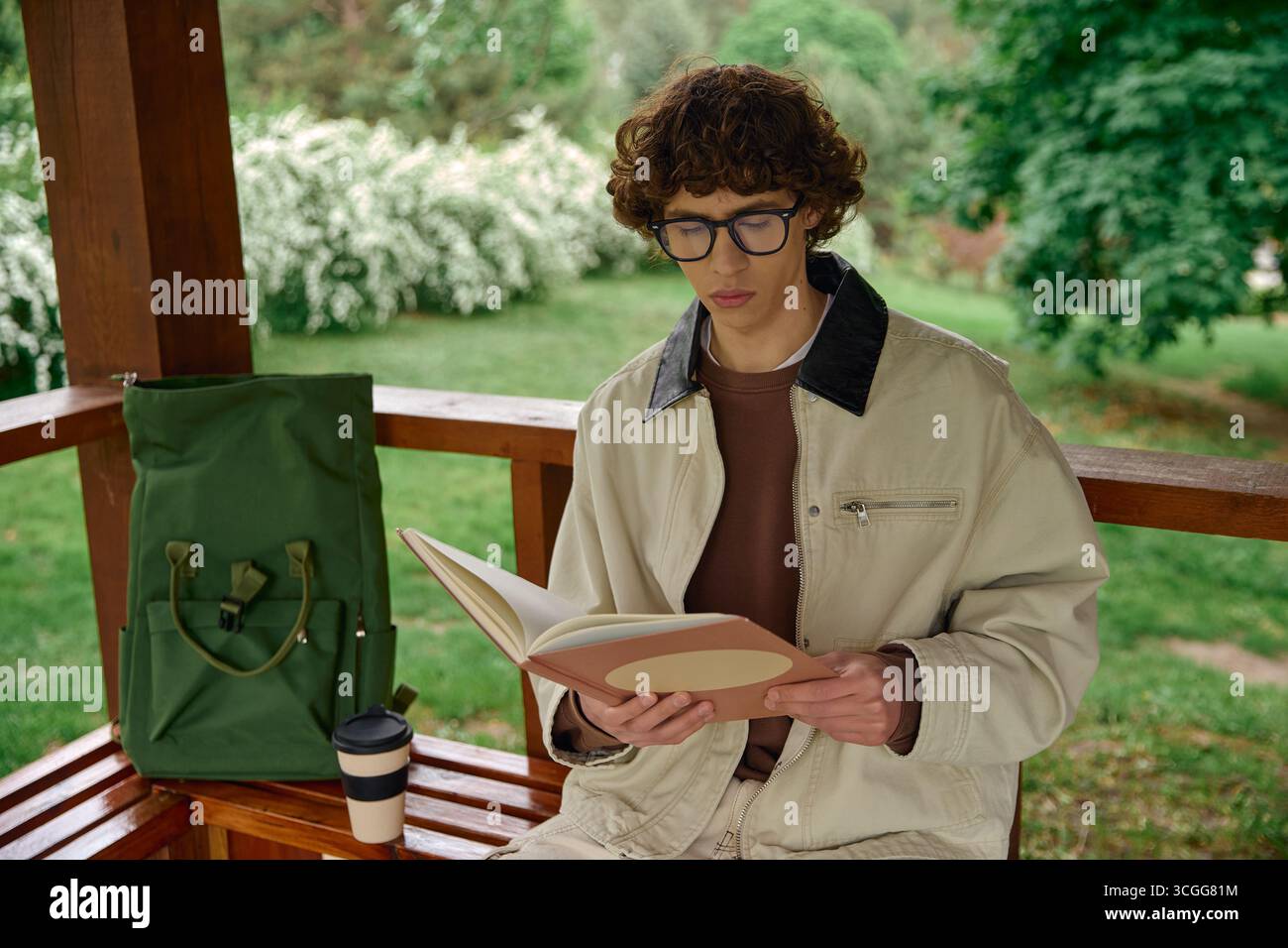 Étudiant se détend à l'extérieur, lisant un livre tout en sirotant un café dans un environnement vert serein. Banque D'Images