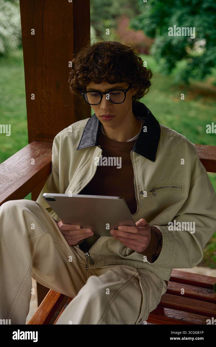 Un jeune homme habillé de façon décontractée est assis dans un cadre paisible en plein air, concentré sur sa tablette. Banque D'Images
