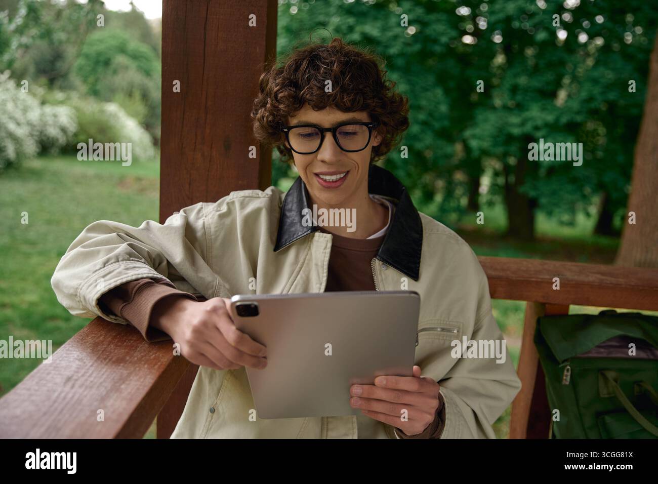 Détendu jeune homme souriant tout en utilisant une tablette sur un porche en bois entouré de verdure. Banque D'Images