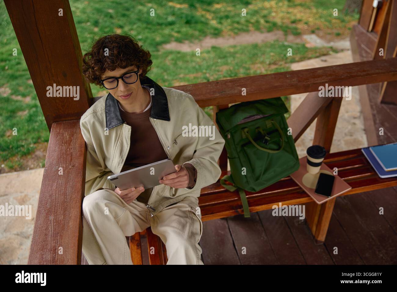 Jeune homme s'engage avec sa tablette tout en se relaxant sur un banc entouré d'une verdure luxuriante. Banque D'Images