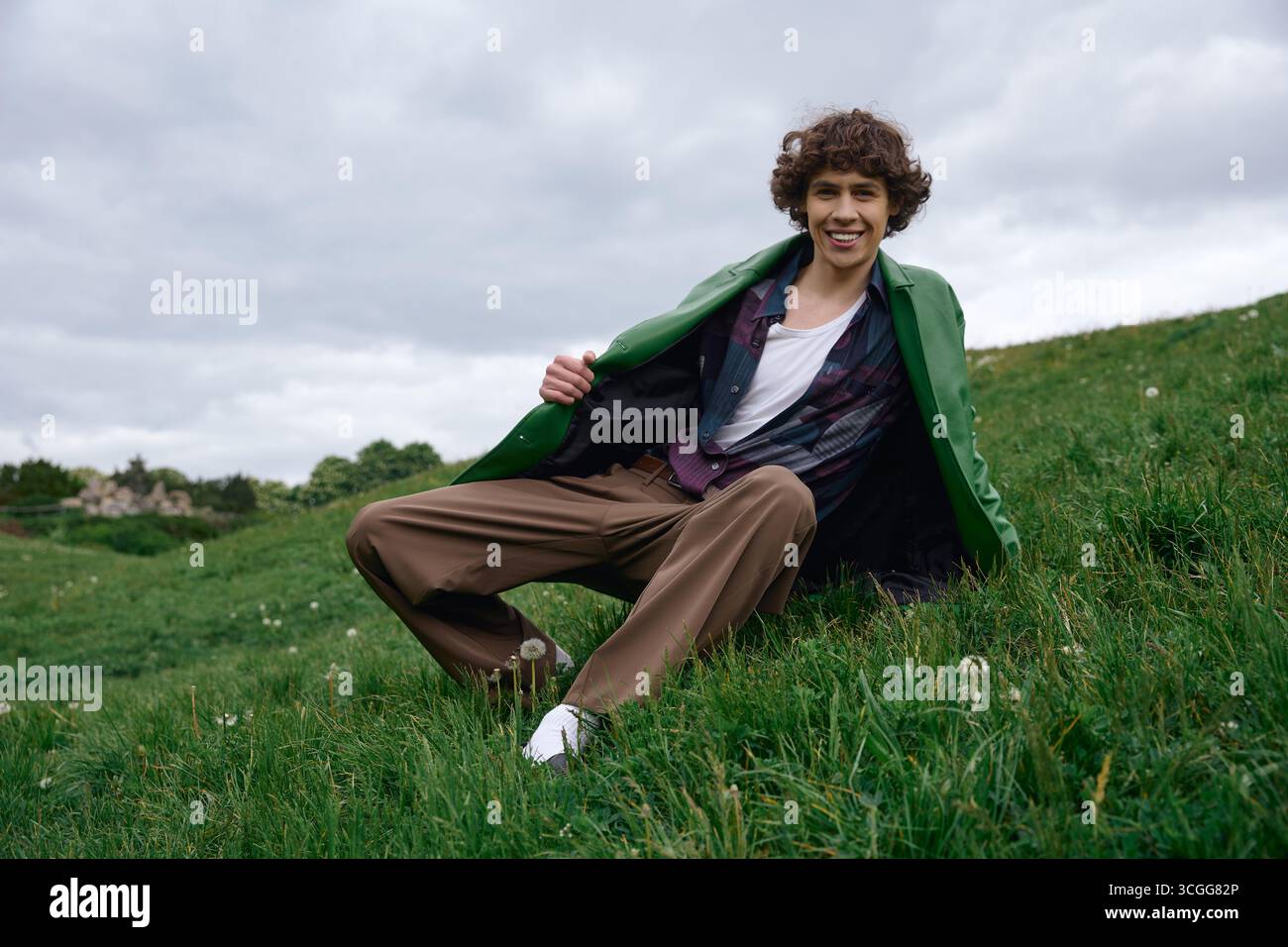 Un jeune homme en tenue décontractée est assis sur l'herbe, souriant tout en profitant d'un moment de paix en plein air. Banque D'Images