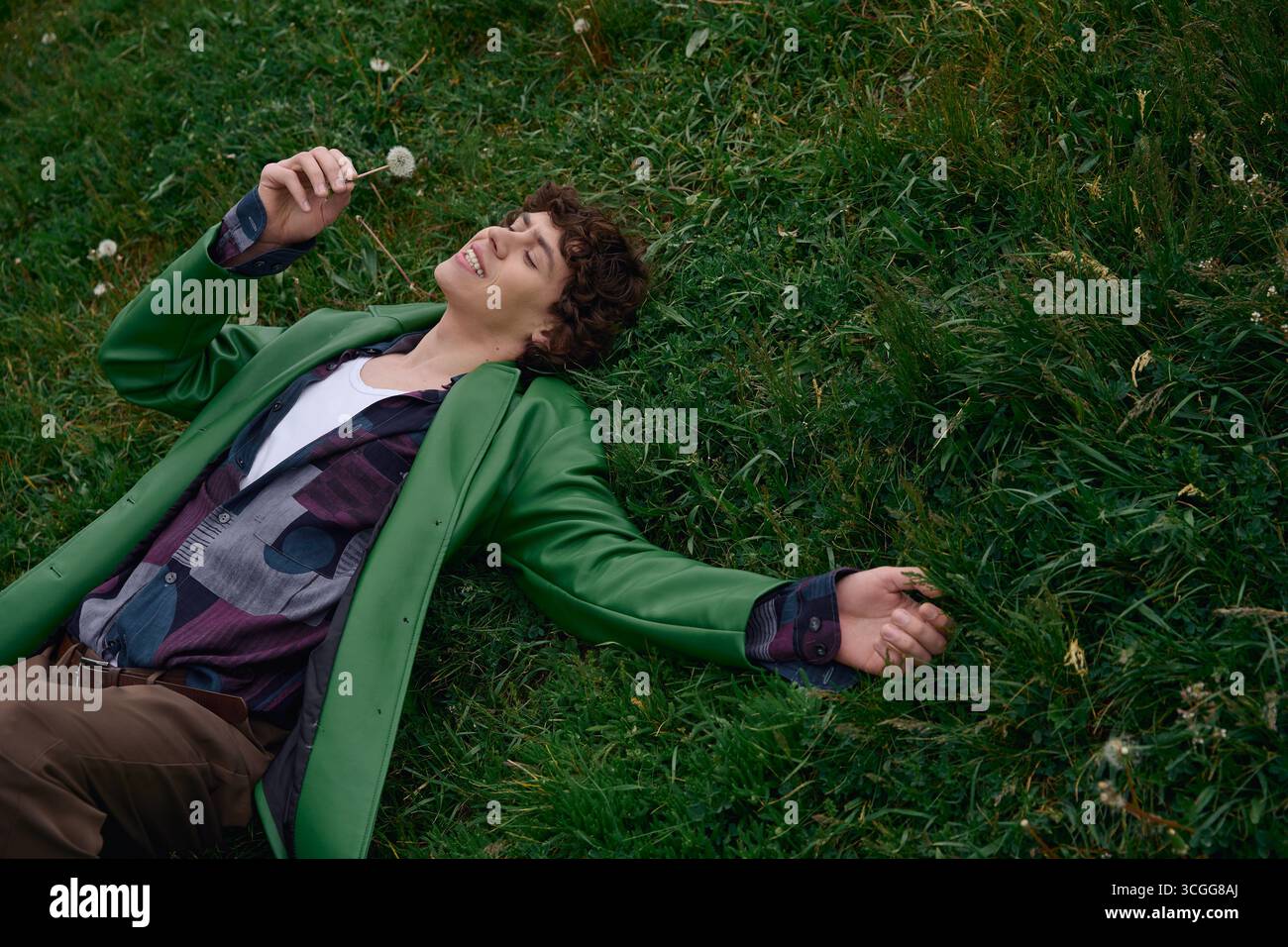 Un jeune homme se détend sur l'herbe, souriant comme il tient un pissenlit dans un cadre extérieur tranquille. Banque D'Images