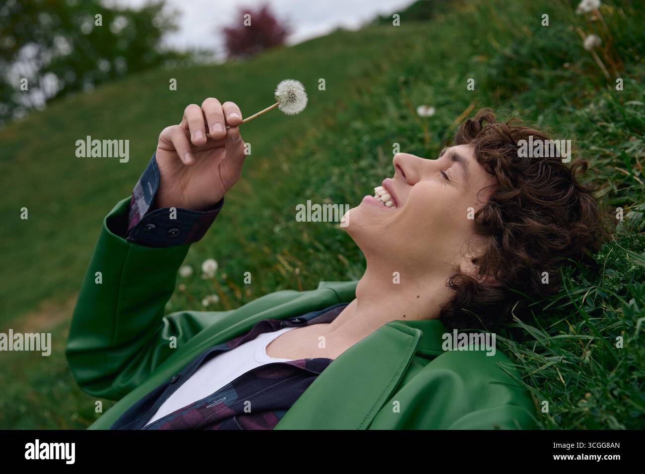 Un bel étudiant se détend sur une colline herbeuse, souriant et tenant une fleur de pissenlit à la main. Banque D'Images