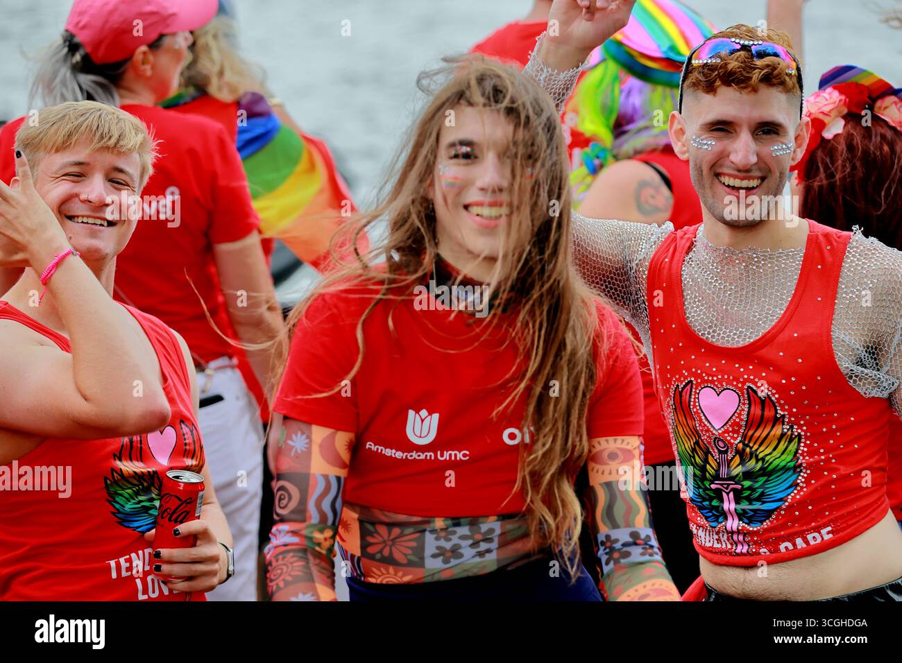 Trois participants souriants en chemises rouges posent sur un bateau Pride Amsterdam canal Parade à Amsterdam, aux pays-Bas, avec de l'eau et un équipage en arrière-plan. Banque D'Images