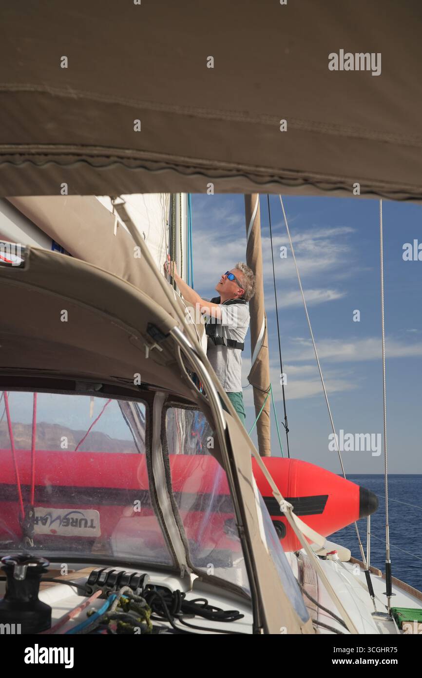 Marin hissant la grande voile au mât sur un yacht de croisière ; bimini et Dodger autour du cockpit avec un gonflable rouge sur le pont, mer calme, lumière du jour Banque D'Images