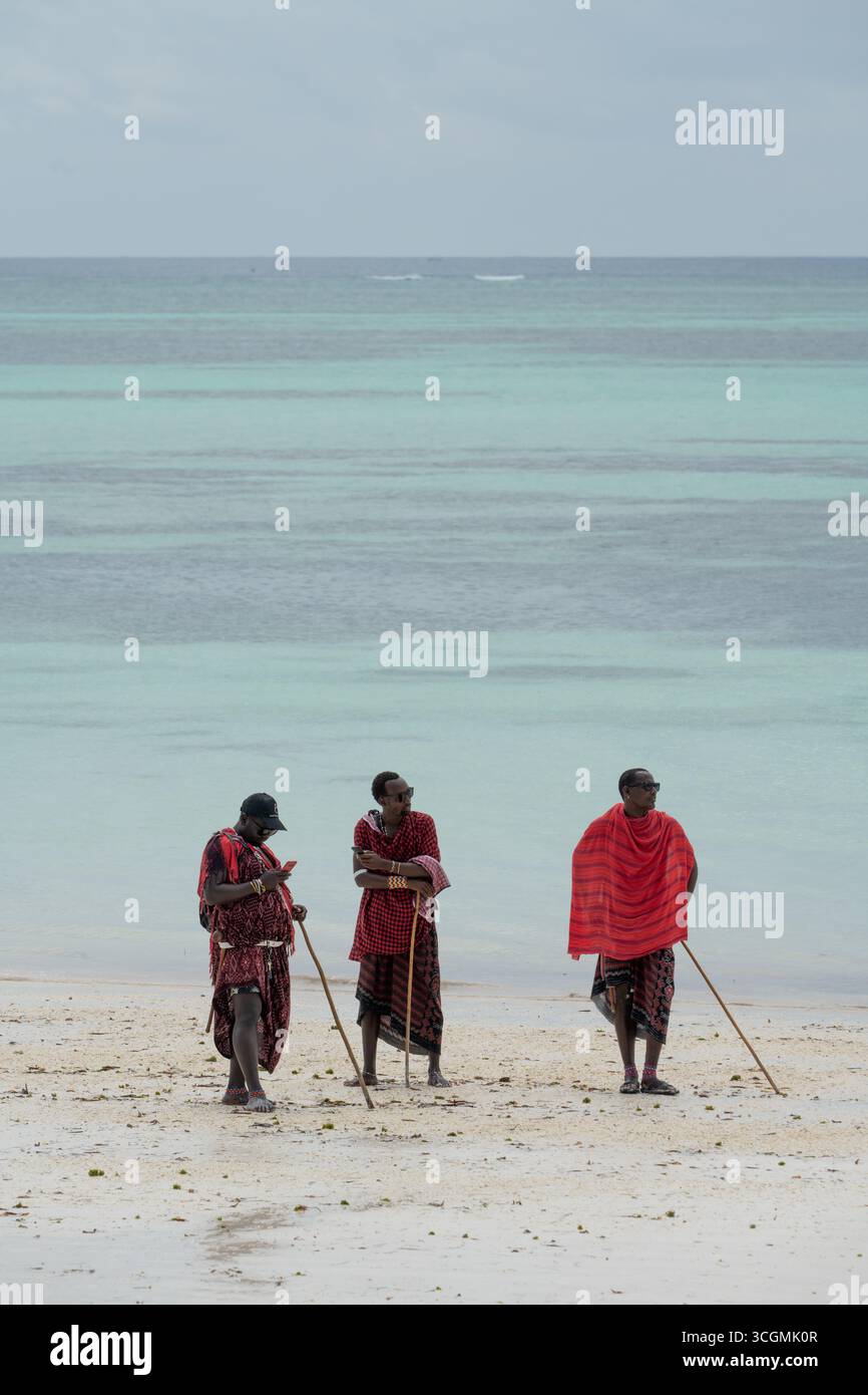 Trois hommes Massaï en robes rouges traditionnelles debout sur la plage, l'un tenant un téléphone portable, montrant la tradition rencontre la vie moderne. Banque D'Images Trois hommes Massaï en robes rouges traditionnelles debout sur la plage, l'un tenant un téléphone portable, montrant la tradition rencontre la vie moderne. Banque D'Images