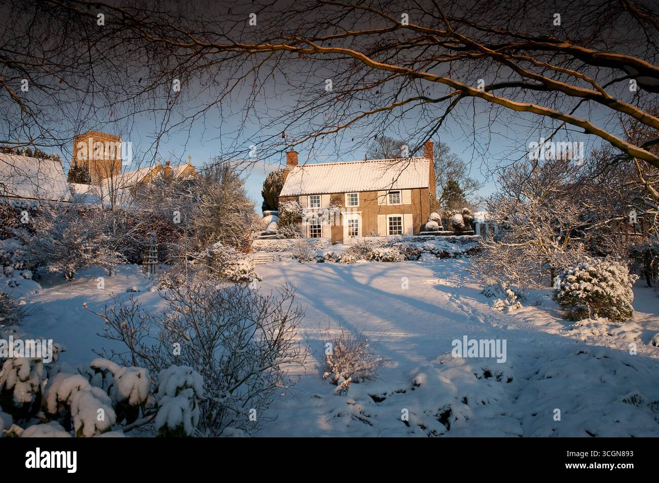 Une charmante maison se trouve au milieu d'une cour enneigée, avec des branches montrant le gel et la lumière douce créant une atmosphère hivernale sereine. Banque D'Images