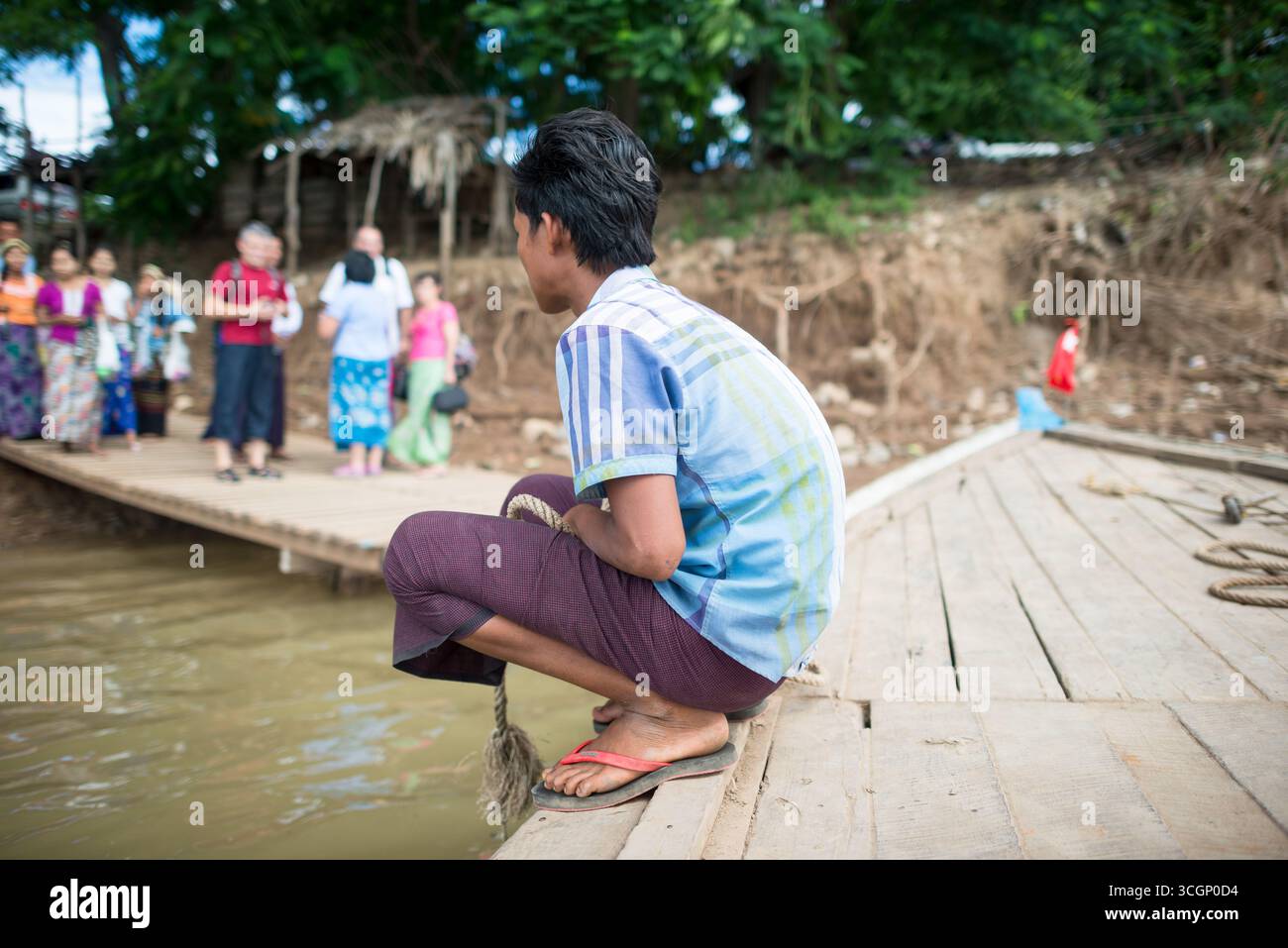 Matelot préparant la corde d'amarrage fleuve Myitnge Amarapura Myanmar | AMARAPURA, Myanmar — Un matelot est accroupi sur une jetée de ferry sur le fleuve Myitnge, une corde à la main, alors que le bateau approche du quai. Le Myitnge, un affluent de rive gauche de l'Ayeyarwady historiquement connu sous le nom de Dokhtawaddy, reste un couloir de travail pour le transport local près d'Amarapura, une ancienne capitale royale dans la région de Mandalay proche de la ville fossée d'Inwa. Banque D'Images