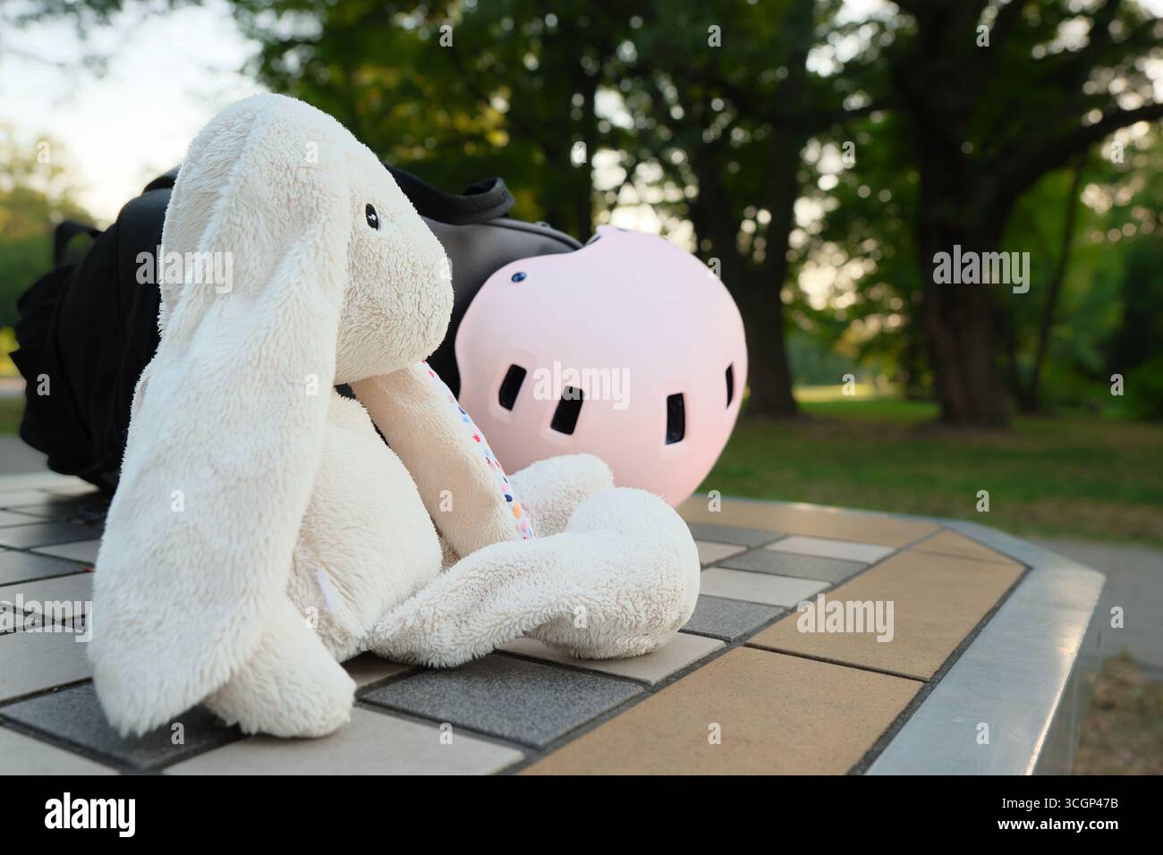 Jouet lapin en peluche avec casque rose et sac à dos sur une table carrelée dans un parc, symbole de la sécurité de l'enfance. Banque D'Images