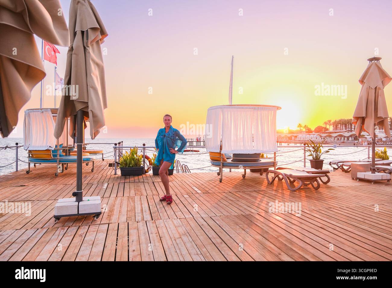 belek, Turkiye, 27.07.2025 Une femme debout sur une jetée en bois au coucher du soleil. Mer calme à l'heure dorée avec fond de station balnéaire. Détente et vacances c Banque D'Images
