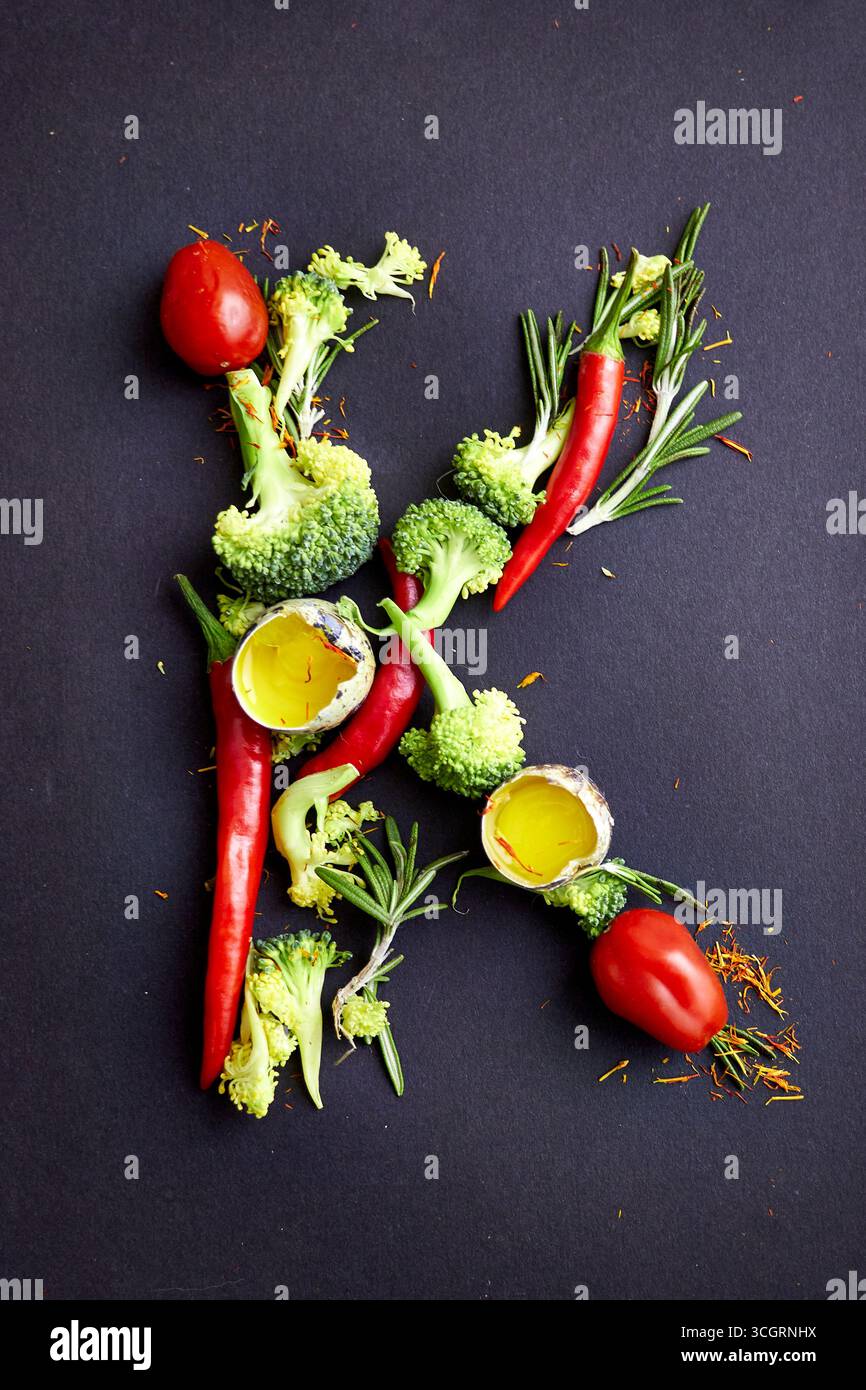 Lettre créative K conçue avec des piments, du brocoli, du romarin, des tomates et de l'huile d'olive dans des coquilles d'oeuf sur fond sombre. Typographie alimentaire unique Banque D'Images