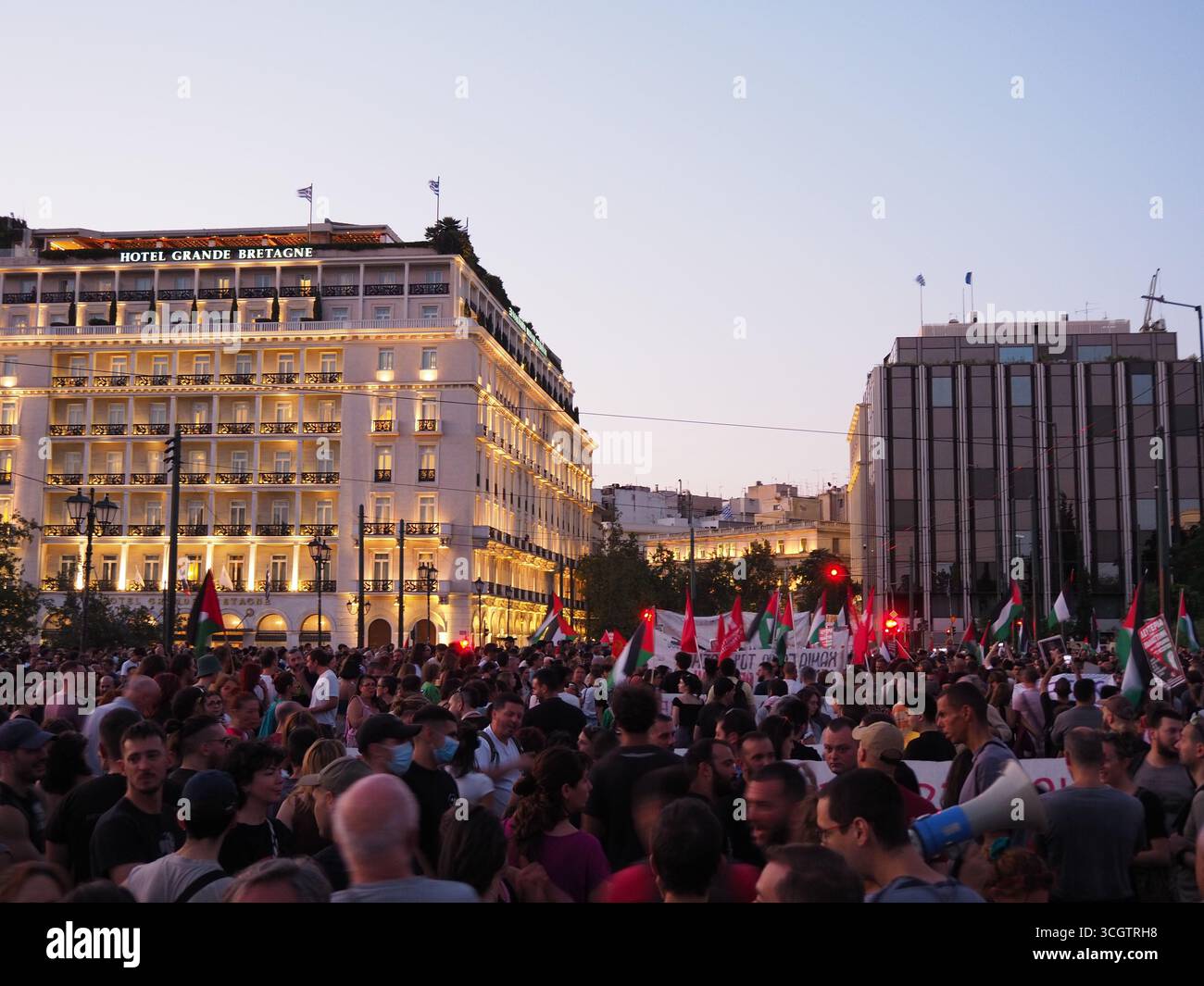 Athènes, Grèce - 10 août 2025 : grande foule avec des drapeaux rouges rassemblés dans le centre-ville d'Athènes au crépuscule, avec des bâtiments illuminés en arrière-plan Banque D'Images