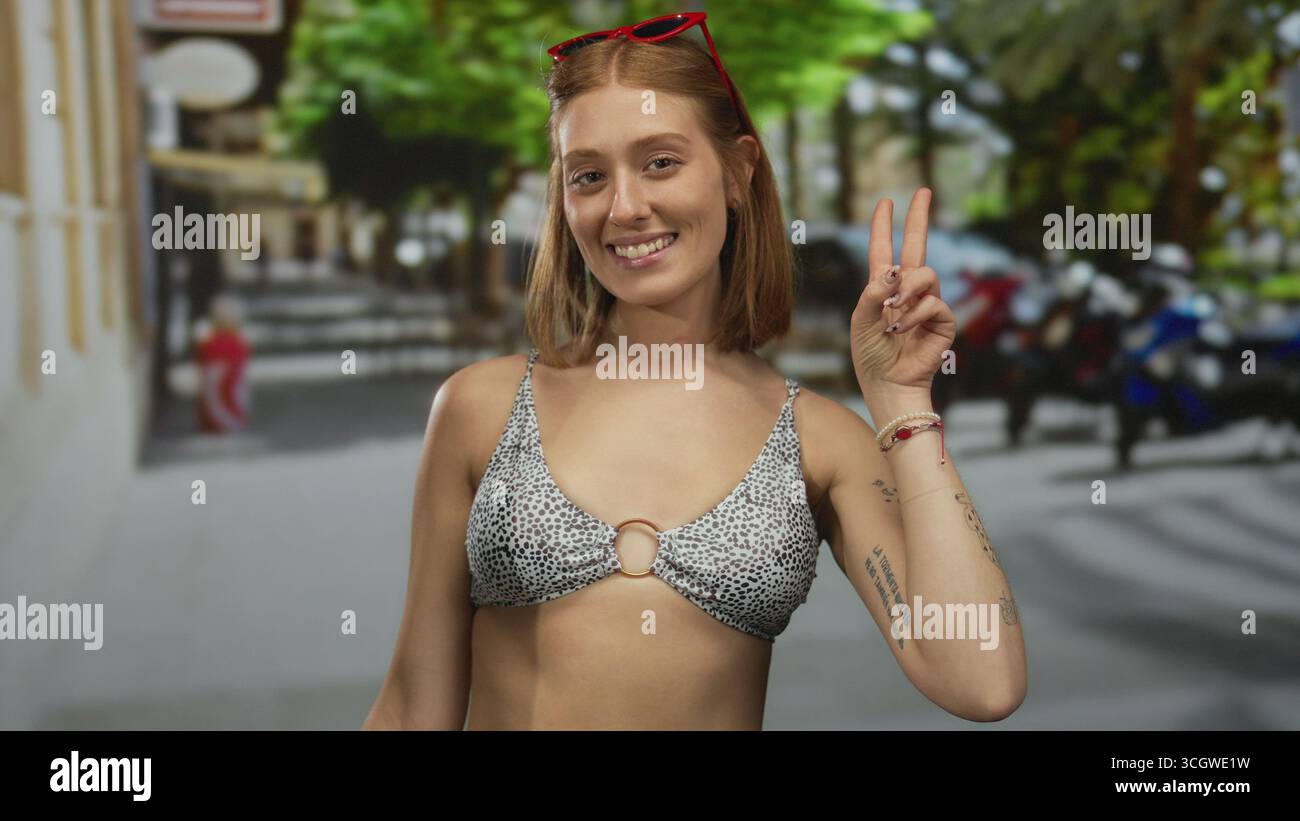 Femme souriant en bikini sur la rue urbaine avec des lunettes de soleil et des tatouages, faisant signe de paix avec fond de nature et accessoires colorés. Banque D'Images
